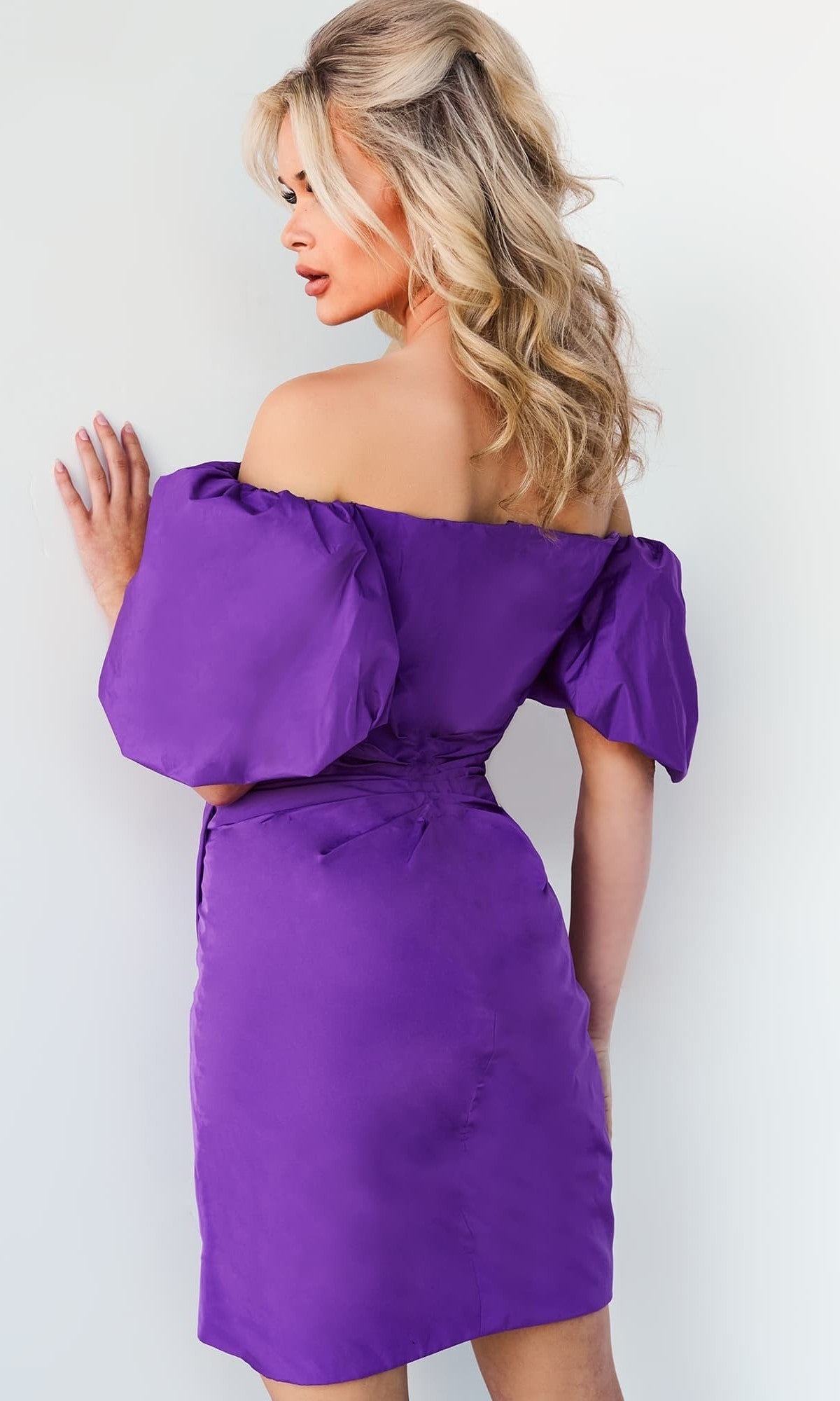 A woman with wavy blonde hair, facing away from the camera against a light background, wears the Jovani 09476 short party dress—an off-the-shoulder purple design with puffed sleeves.